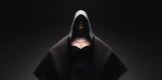 Model in dramatic oversized cocooning coat on a pitch-black runway with chiaroscuro spotlight lighting representing the Balenciaga runway 2026 ClairObscur collection by Pierpaolo Piccioli at Paris Fashion Week