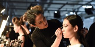 Makeup artist applying editorial runway beauty look to a model backstage at a fashion week show, representing the Beauty Trends Hub covering runway makeup, hair, and skincare trends