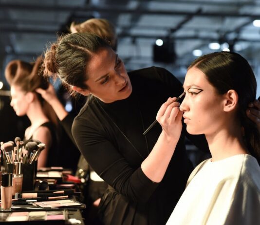 Makeup artist applying editorial runway beauty look to a model backstage at a fashion week show, representing the Beauty Trends Hub covering runway makeup, hair, and skincare trends