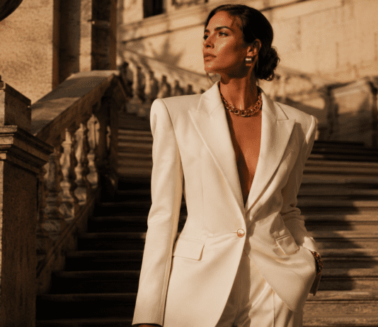 Model in a structured ivory wide-shoulder blazer with gold accessories on a sunlit stone staircase representing the new maximalism fashion trend in spring summer 2026