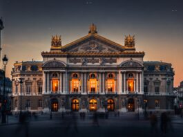 The Palais Garnier opera house in Paris at dusk, golden interior light through arched windows, the ornate neoclassical facade representing the Paris Opera Ballet's 2026 season at the center of a global classical versus contemporary ballet debate.
