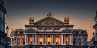 The Palais Garnier opera house in Paris at dusk, golden interior light through arched windows, the ornate neoclassical facade representing the Paris Opera Ballet's 2026 season at the center of a global classical versus contemporary ballet debate.