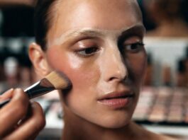 Model's face mid-underpainting makeup application with cream contour blended along the cheekbone before a sheer foundation layer is applied, demonstrating the reverse contouring technique used by professional makeup artists.
