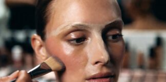 Model's face mid-underpainting makeup application with cream contour blended along the cheekbone before a sheer foundation layer is applied, demonstrating the reverse contouring technique used by professional makeup artists.
