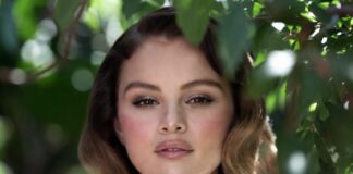 Woman in white tulle dress in a lush garden with rosy pink blush shimmering eyeshadow ultra-glossy lips and soft waves representing the Selena Gomez Rare Beauty fairy makeup aesthetic and garden photoshoot beauty look from the June 2026 campaign